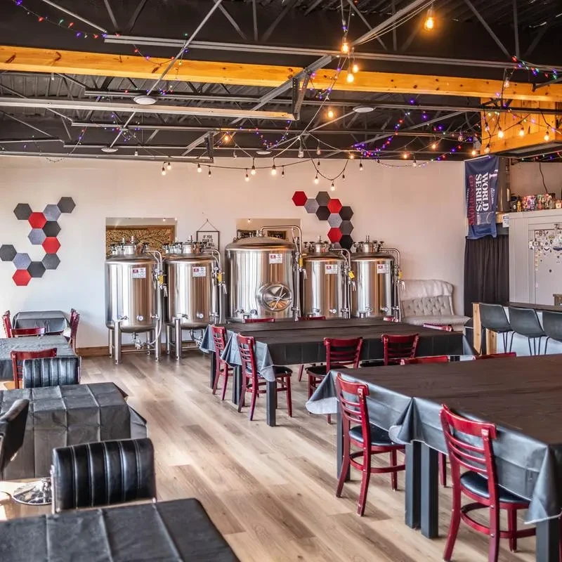 Brewery interior showing four stainless steel tanks, tables, and string lights.