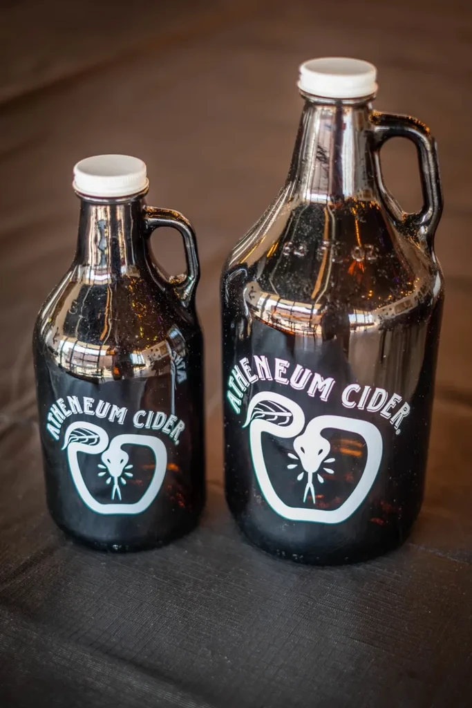 Two dark glass Atheneum Cider growlers, different sizes, snake logo.