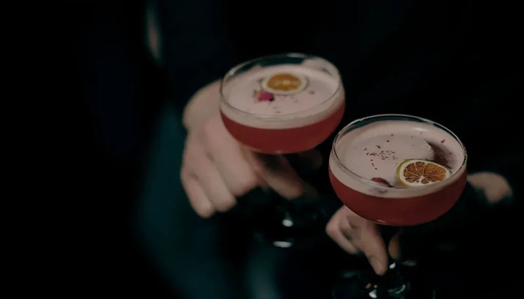 Two vibrant reddish-pink cocktails in stemmed glasses held by hands.