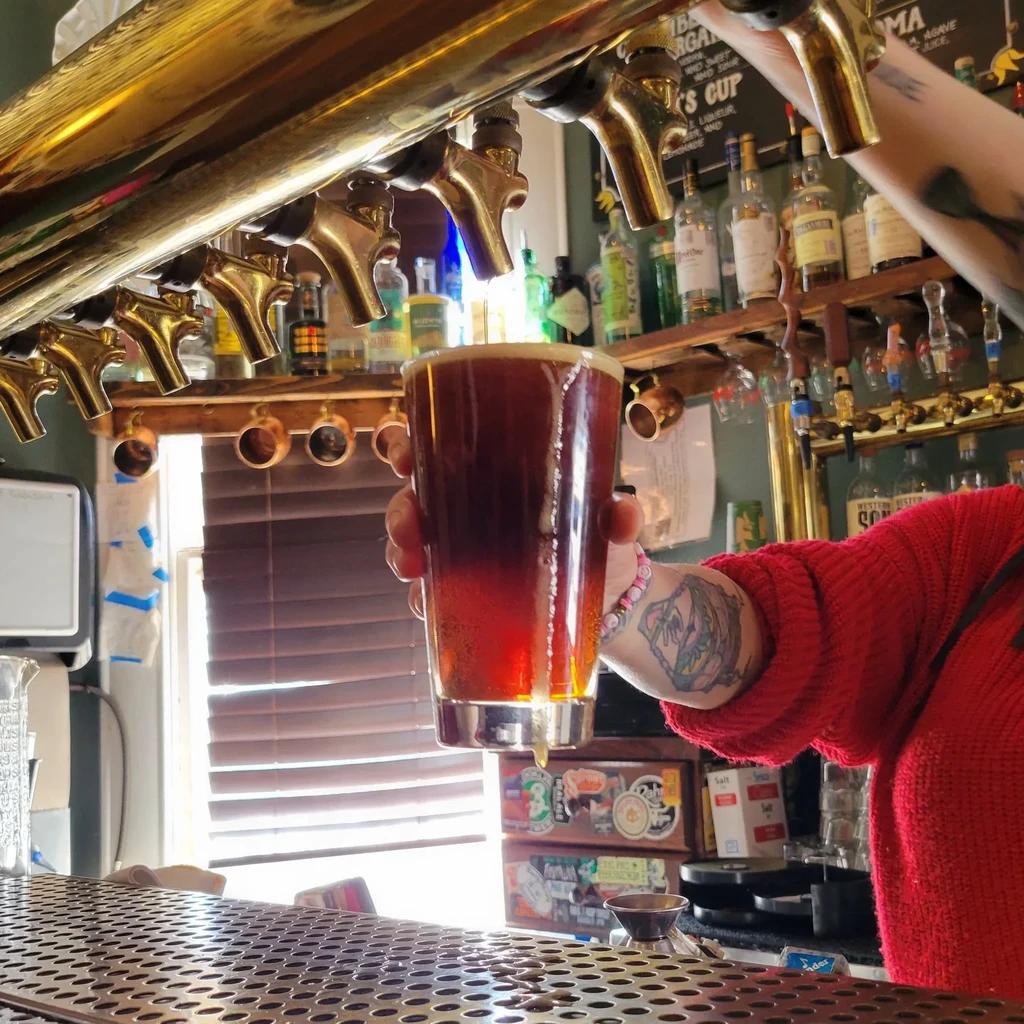 Red-sleeved hand holds a dripping dark beer pint at a bright bar.