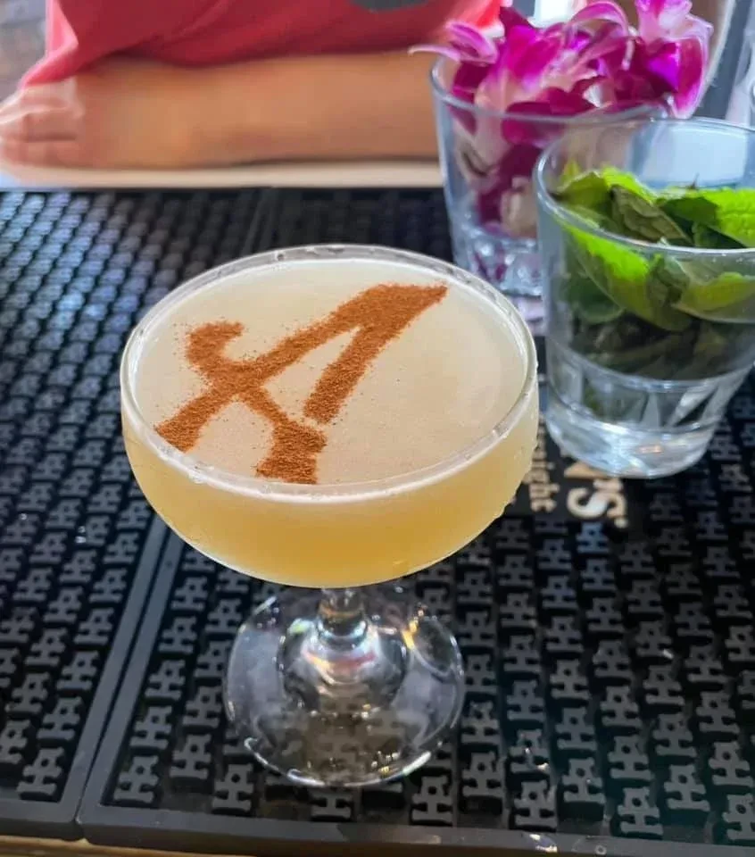 Coupe glass cocktail, pale yellow, with a spiced 'A' on frothy top.