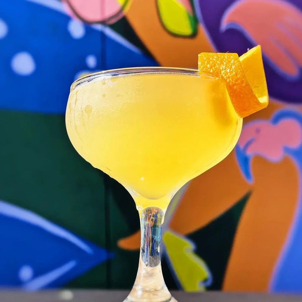 Vibrant yellow cocktail in coupe glass with orange peel, abstract colorful background.