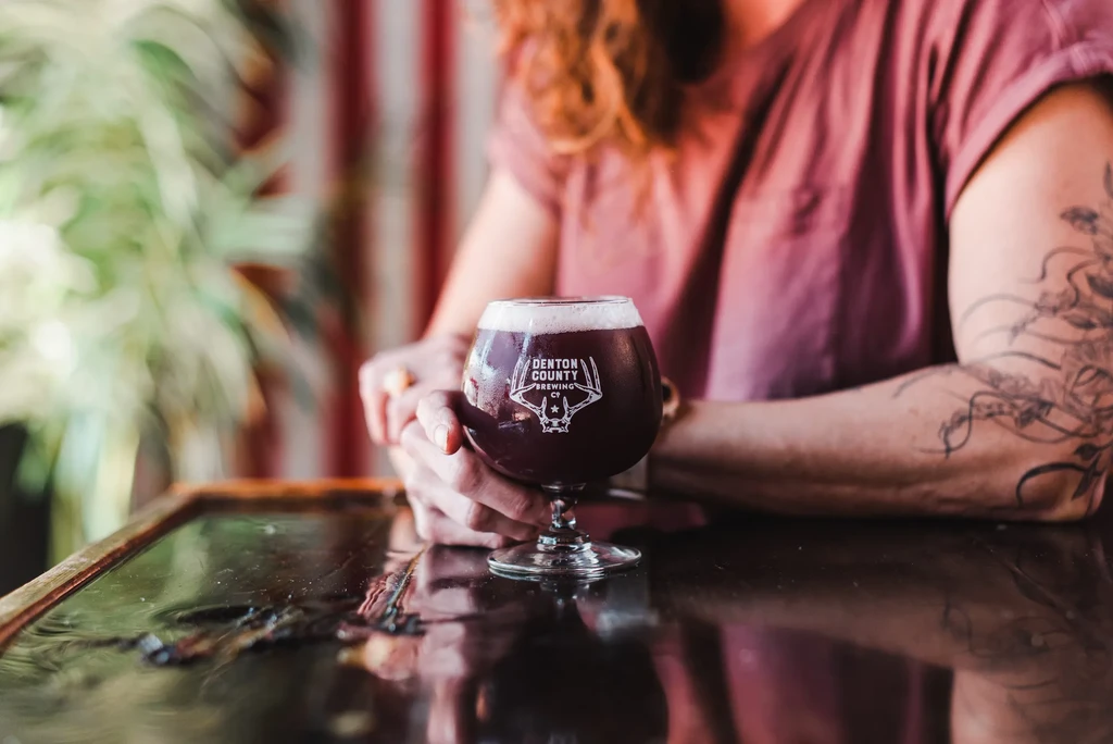 Dark red foamy drink in a stemmed Denton County Brewing Co glass.