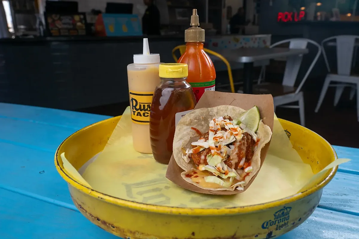 Crispy chicken taco with slaw on yellow tray, sauces, blue table