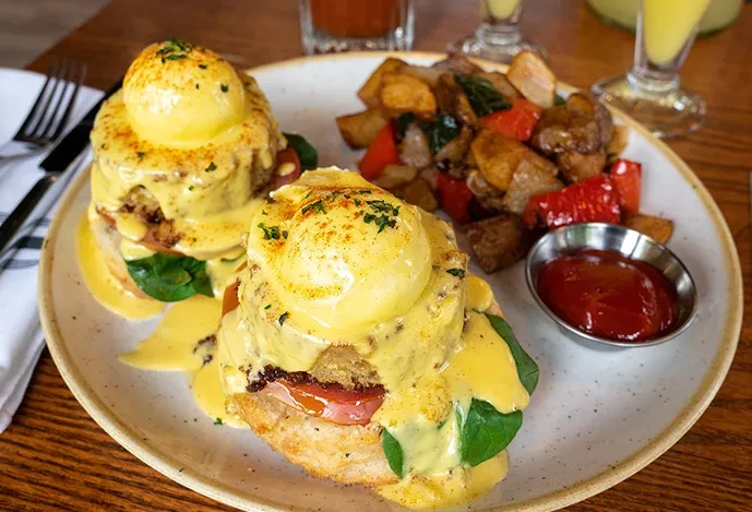 Close-up: Two Eggs Benedict, roasted potatoes, and sauces on a plate.