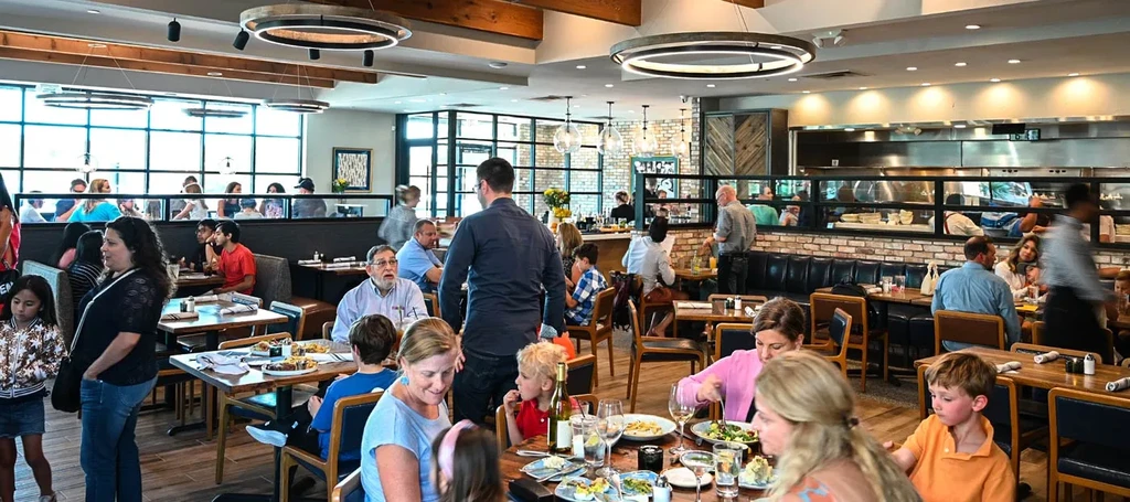 Bustling modern-rustic restaurant interior with patrons and visible open kitchen.