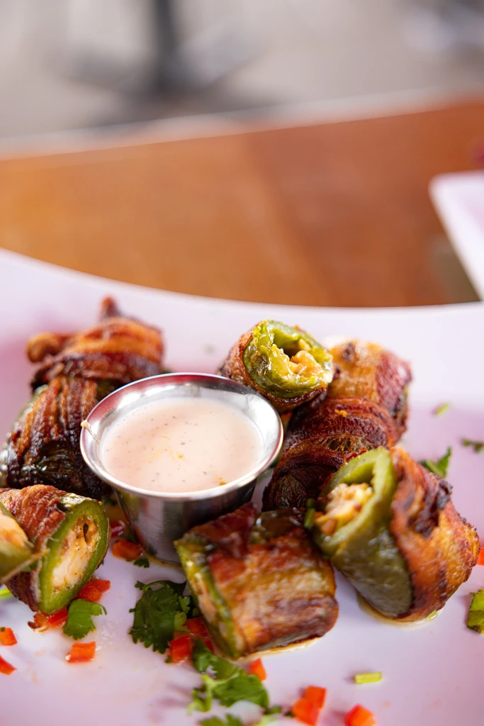 Golden bacon-wrapped jalapeño poppers with dipping sauce, garnished with cilantro.
