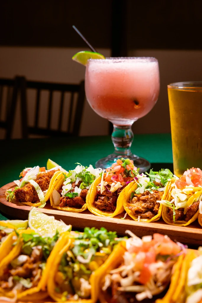 Six colorful tacos with various toppings and drinks on a wooden board.