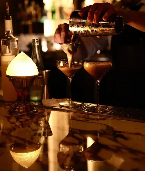 Bartender strains cocktail into coupe glass at a dimly lit bar.