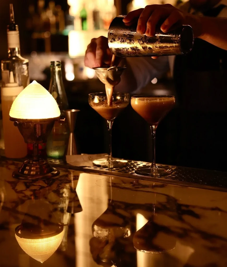 Bartender's hands strain reddish cocktail from shaker into two coupe glasses.