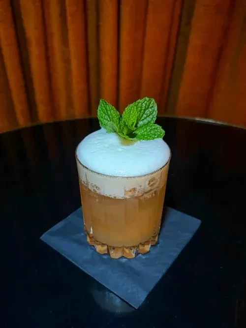 Amber cocktail with white foam and green mint in faceted glass.
