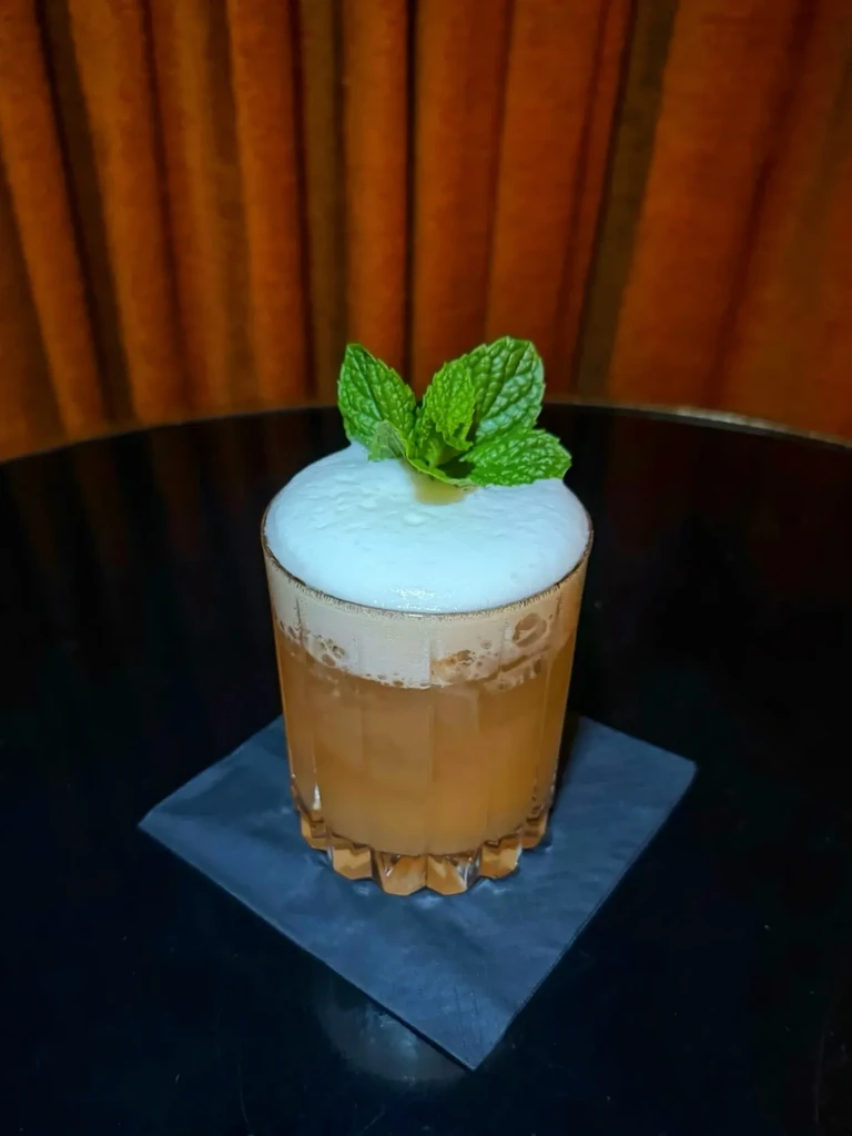 Faceted glass of amber cocktail, frothy foam, and mint garnish.