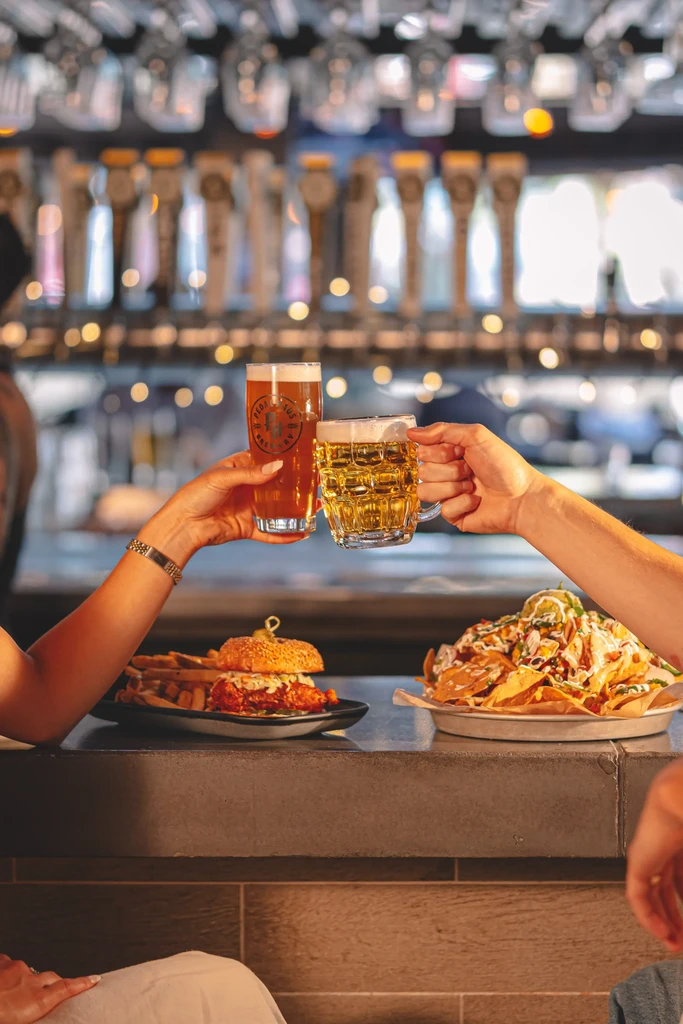 Two hands clinking beers, with fried chicken and loaded nachos.