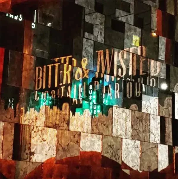 Illuminated "Bitter & Twisted Cocktail Parlor" sign on a textured wall with green neon.