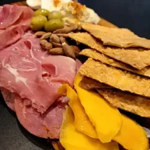 Charcuterie board with meats, olives, almonds, creamy dip, crackers, and mango.