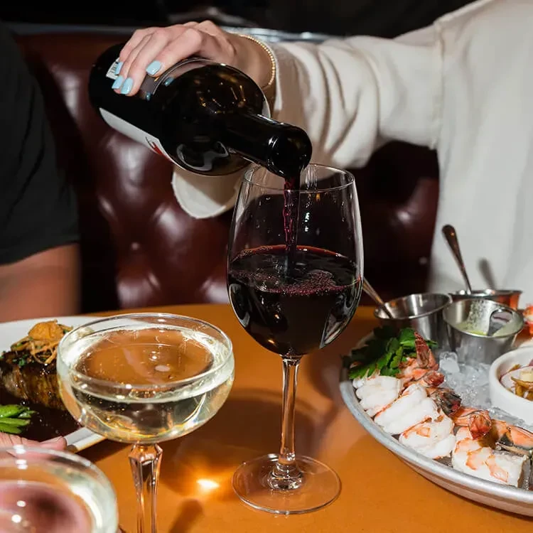 Hand with blue nails pours red wine; shrimp cocktail, drinks on table.