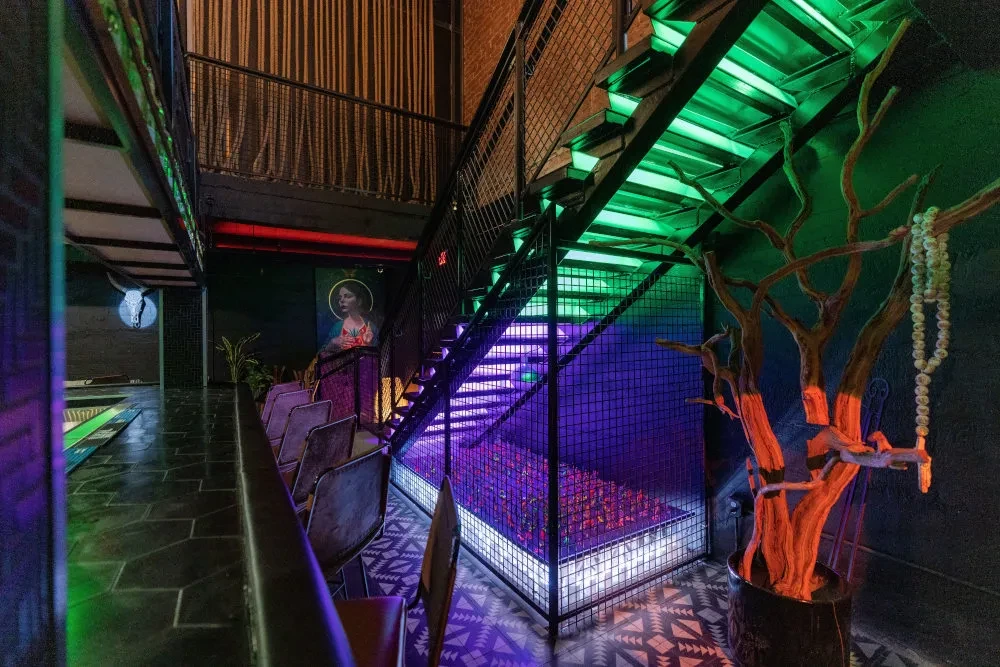 Modern restaurant interior with dramatic green/purple lit staircase and eclectic decor.