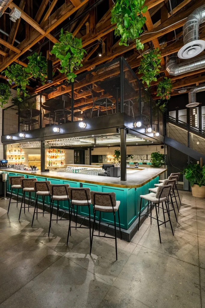 Modern bar with teal counter, mezzanine, exposed beams, and hanging plants.
