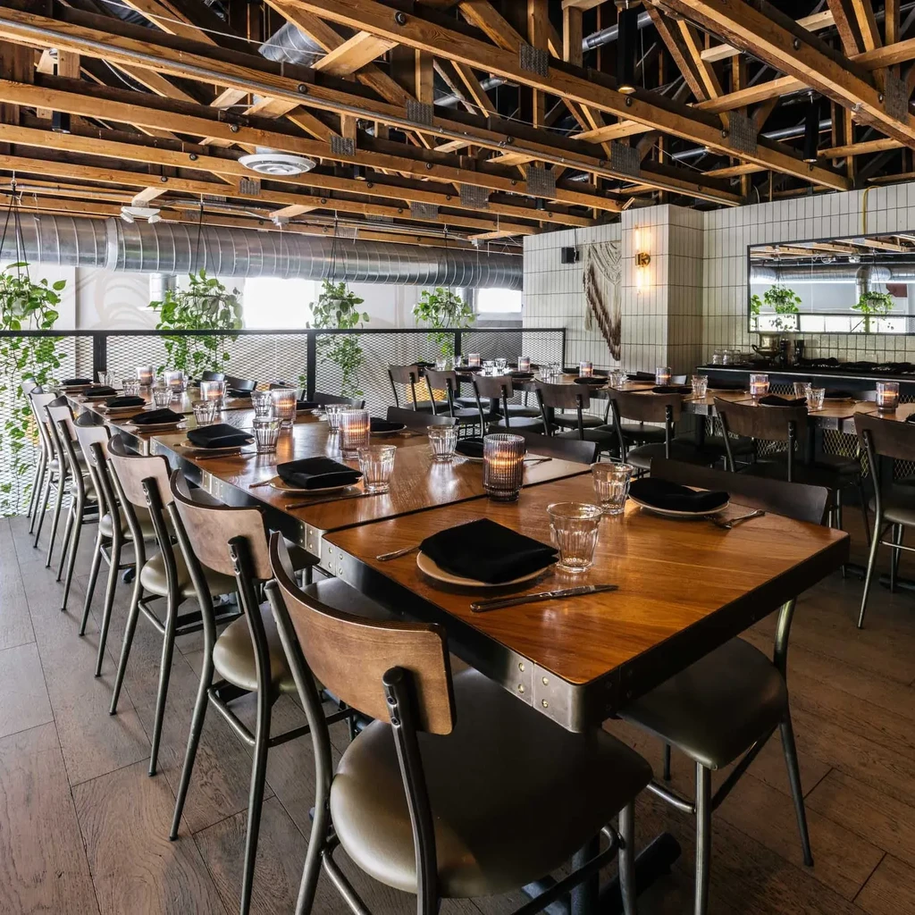 Modern restaurant with long wooden table, industrial ceiling, and plants.