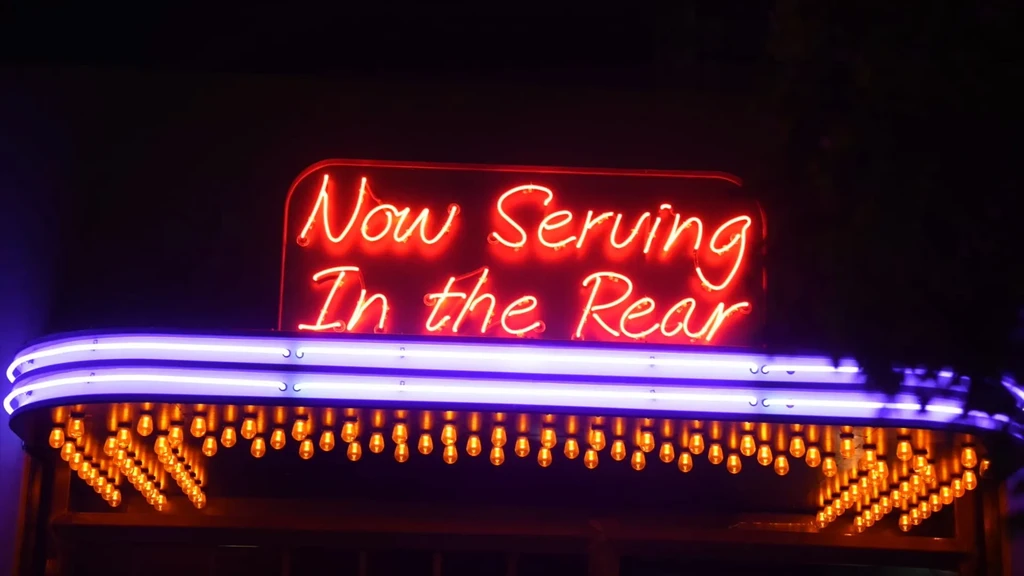 Red neon sign "Now Serving In the Rear" illuminates ornate marquee.