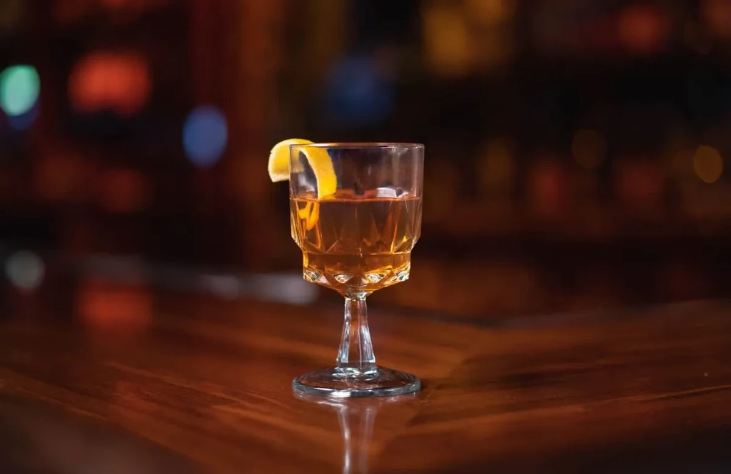 Faceted glass of amber cocktail with lemon peel on dark wood.