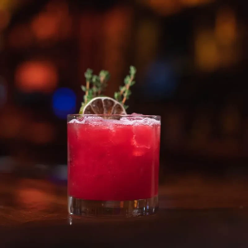 Reddish-pink cocktail in a short glass with dried citrus and green herbs.