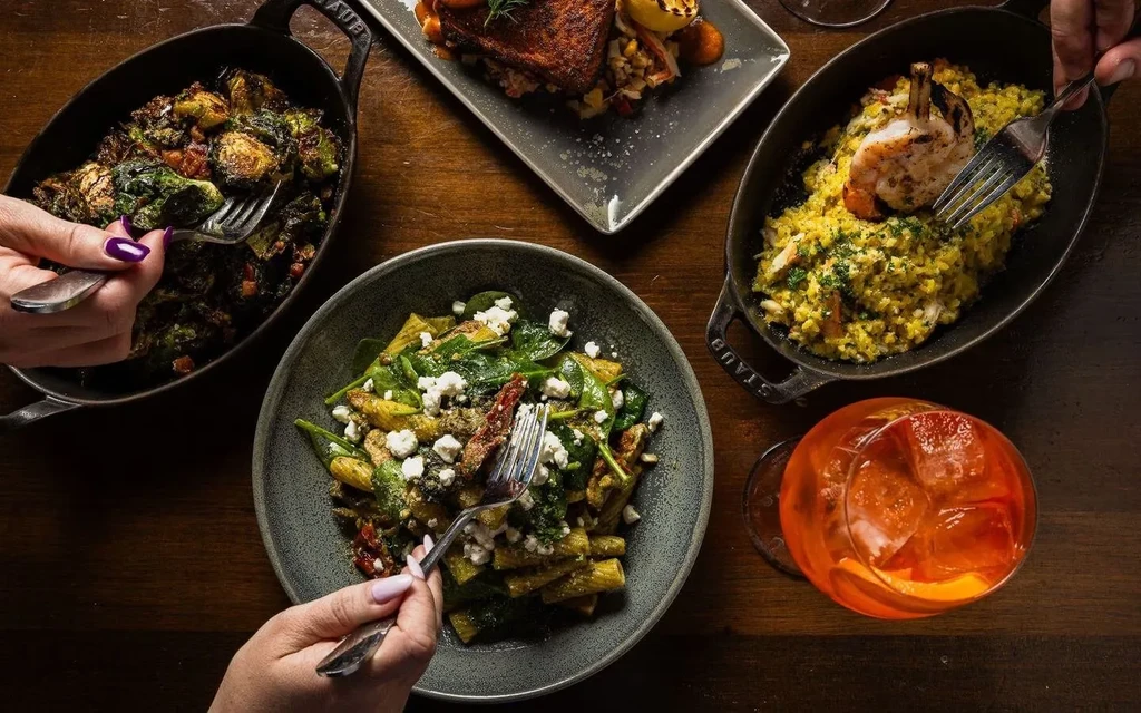 Overhead: Dining table with various dishes, a cocktail, and three hands.