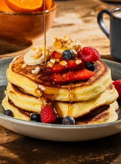 Golden pancakes drizzled with syrup, topped with fresh berries, banana, and nuts.