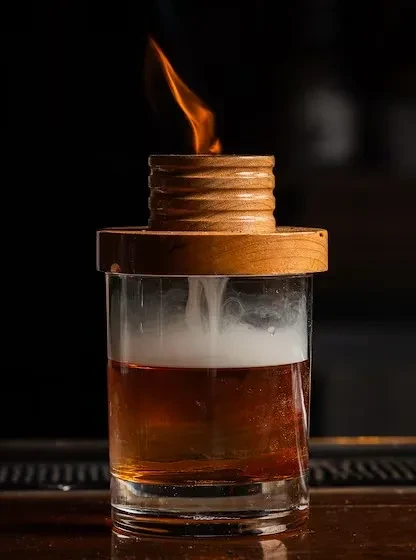 Wooden smoker device burning over amber cocktail with white vapor.