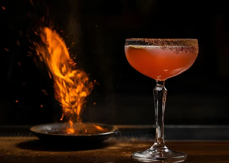 Reddish-orange cocktail with spiced rim next to a flaming dish.