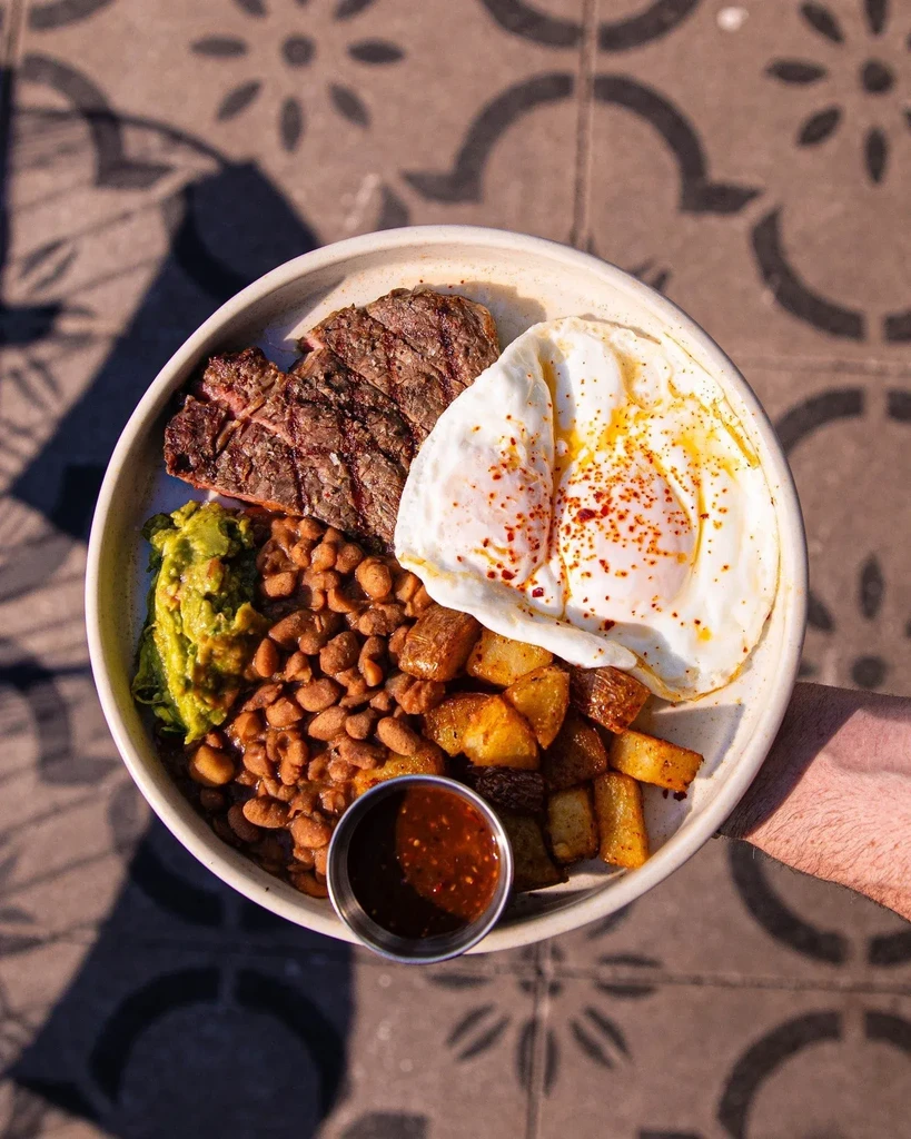 Bowl of grilled steak, fried eggs, beans, guacamole, and potatoes.
