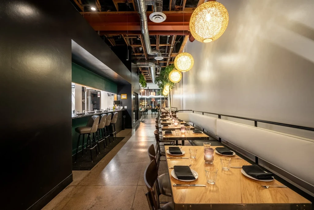Modern restaurant interior with bar, dining tables, industrial ceiling, and foliage.