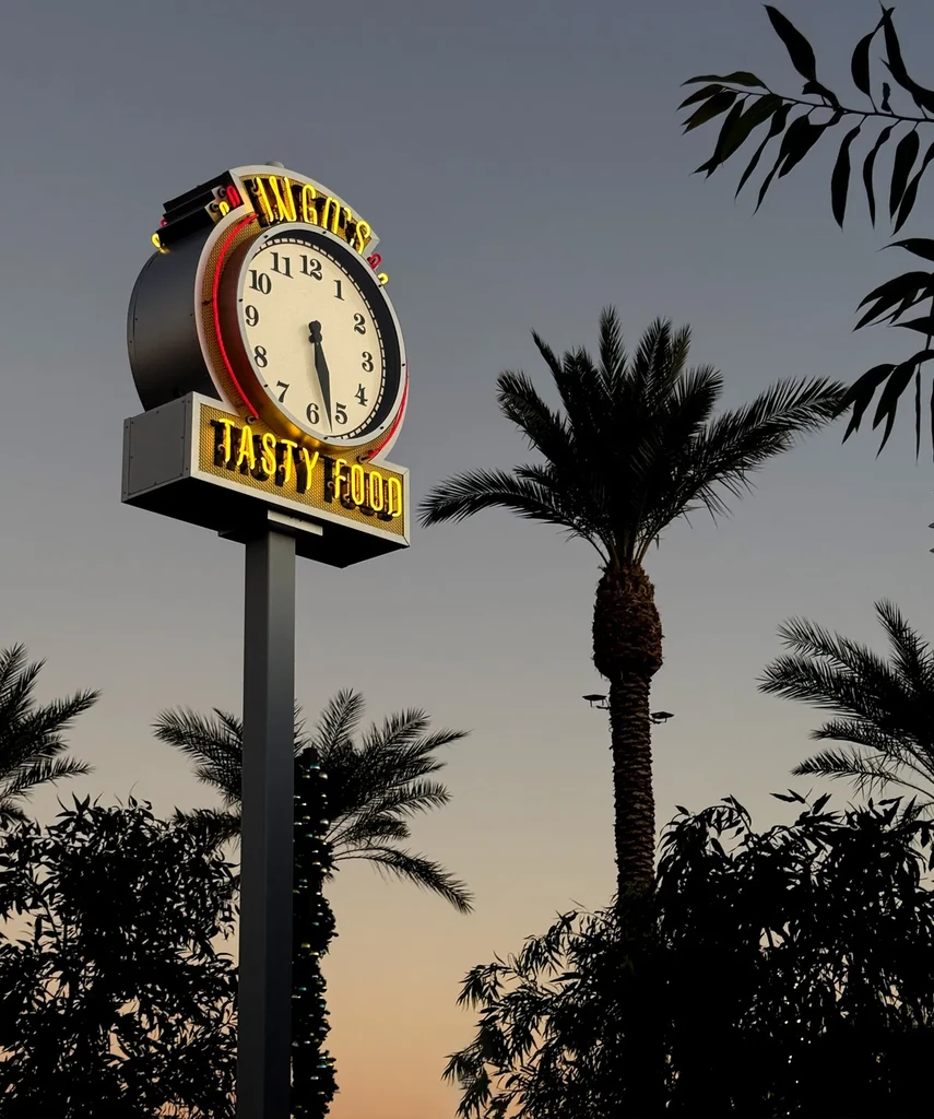 Illuminated Angie's Tasty Food sign with a clock, palm trees at dusk.