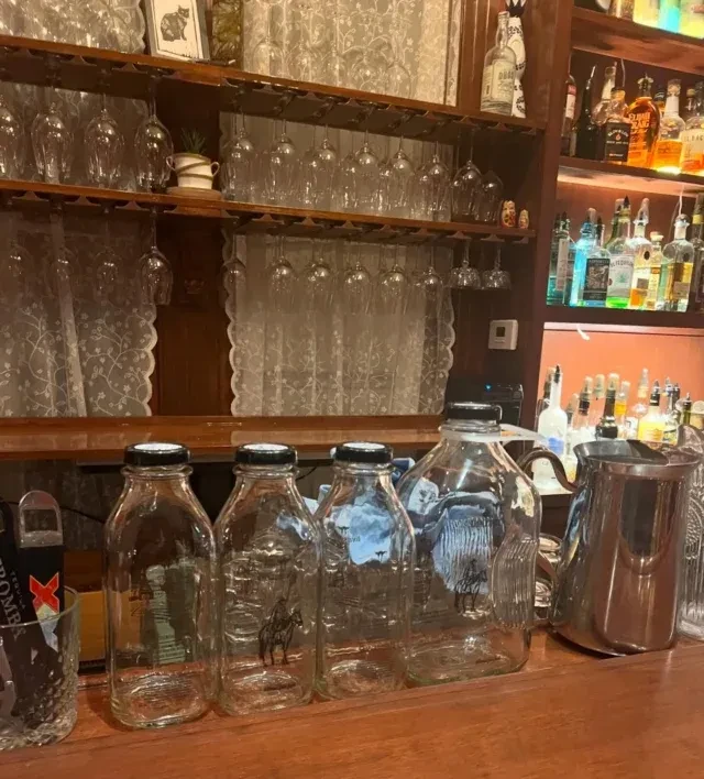 Bar counter with empty bottles, liquor cabinet, wine glasses, and lace curtains.