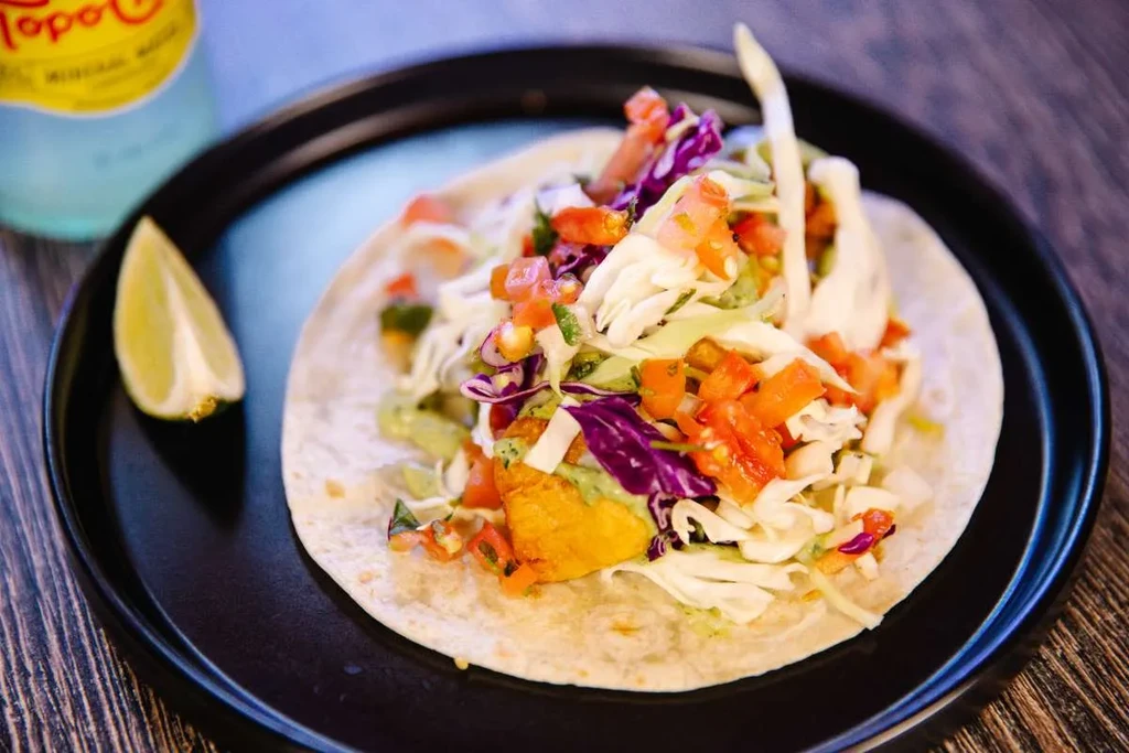 Vibrant fish taco topped with cabbage, tomatoes, sauce, and a lime wedge.