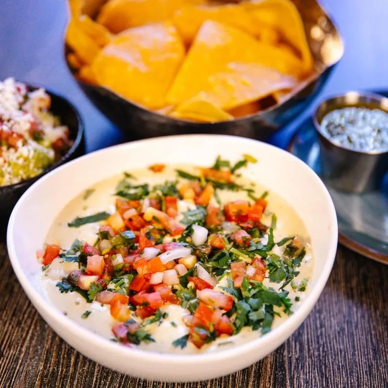 Queso, tortilla chips, guacamole, and sauce on a dark wooden table.