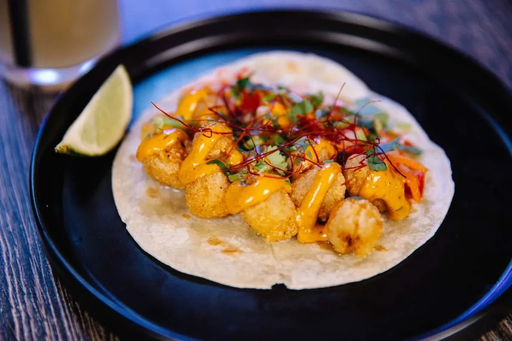 Single soft taco with golden fried filling, orange sauce, herbs, and lime.