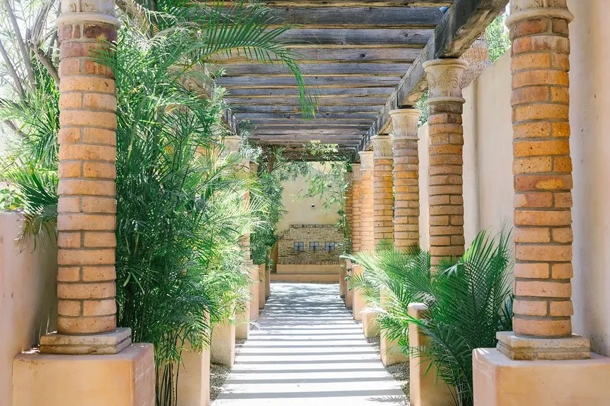 Shaded outdoor path with ornate brick columns and lush palm plants.