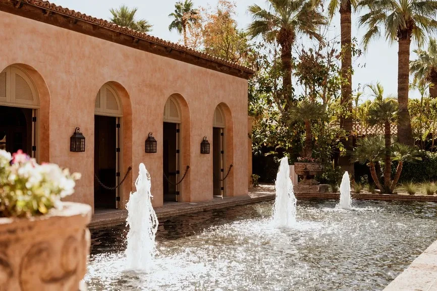 Peach stucco building, arched doorways, fountains, palm trees, and flowers.