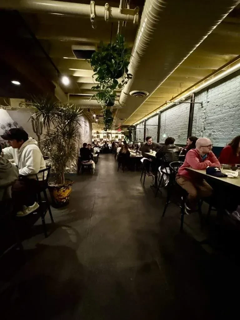 Bustling restaurant with exposed brick, pipes, plants, and warm lighting.