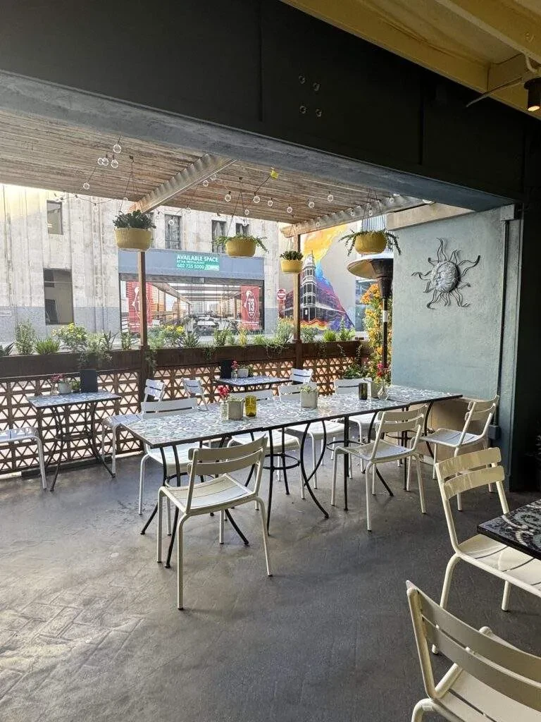 Outdoor restaurant patio with patterned tables, hanging plants, city view.