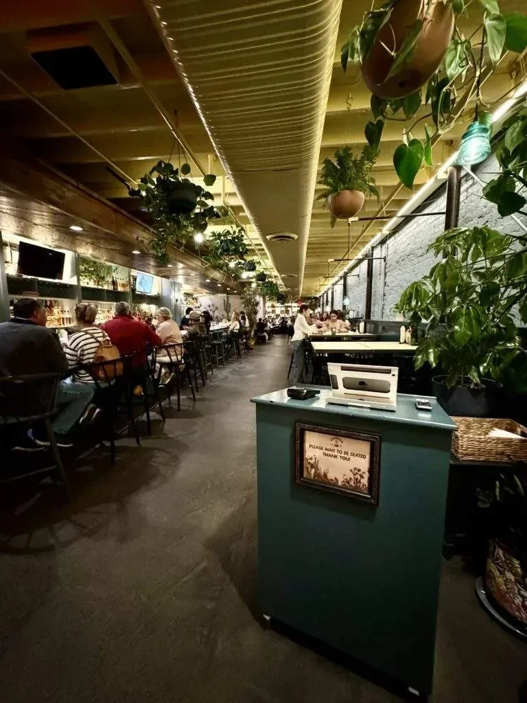 Warm restaurant interior with bar, hanging plants, and host stand.