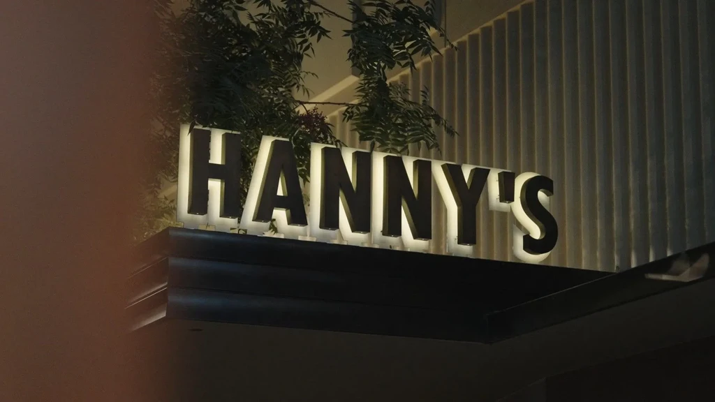 Illuminated 'HANNY'S' sign at night, with green foliage above.