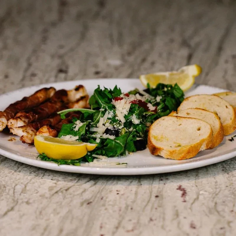 Grilled bacon-wrapped meat, arugula salad, lemon, and crusty bread on a plate.