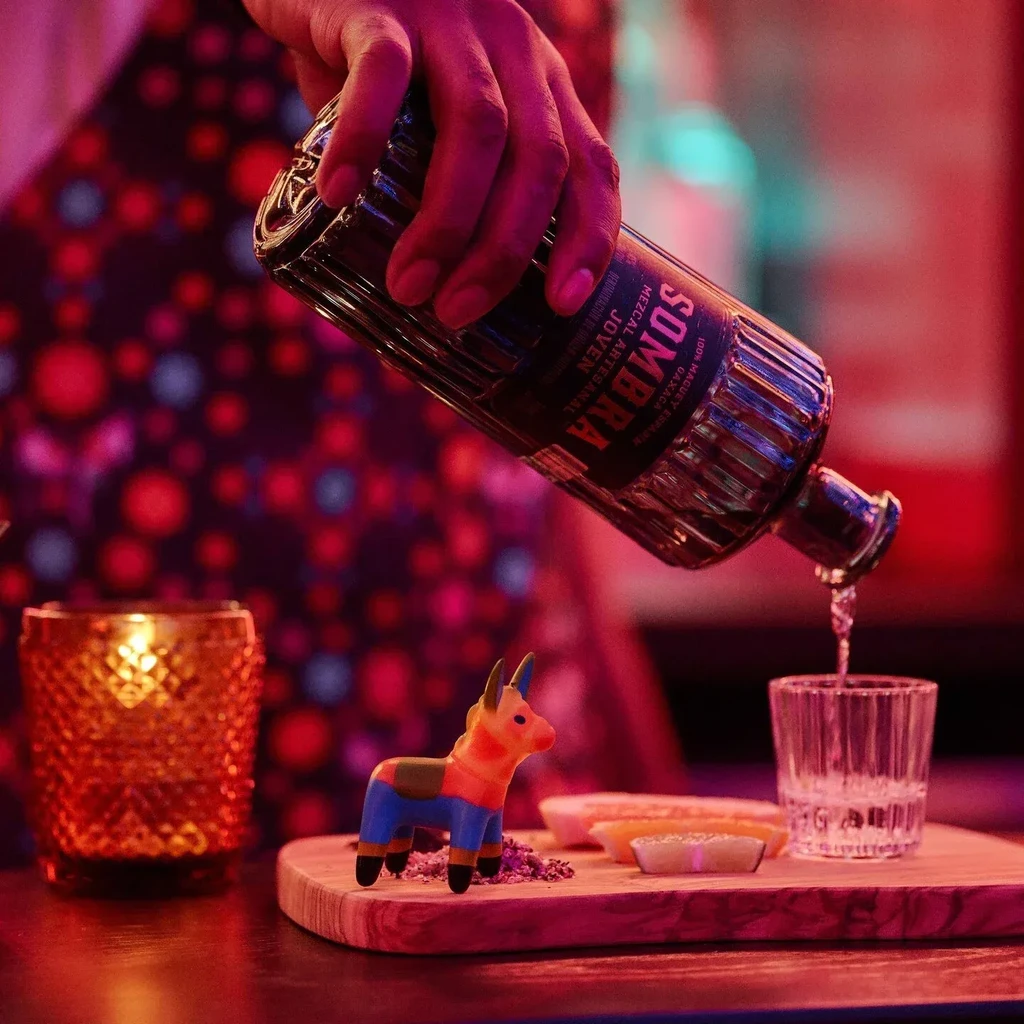 Hand pouring Sombra Mezcal into a shot glass, piñata nearby.