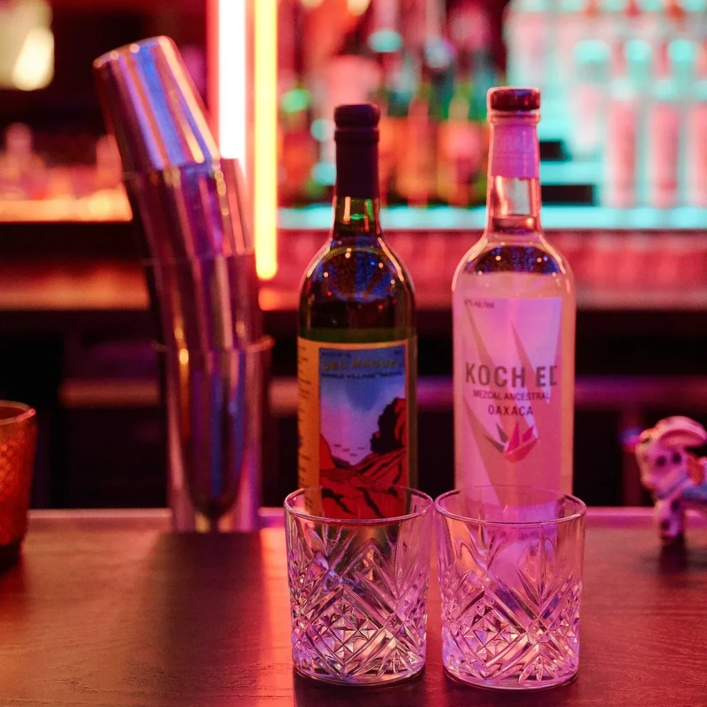 Mezcal bottles, crystal glasses, and cocktail shakers on a neon-lit bar.