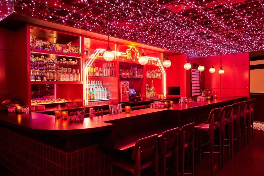 Red-lit bar interior, long counter, liquor shelves, and string lights.