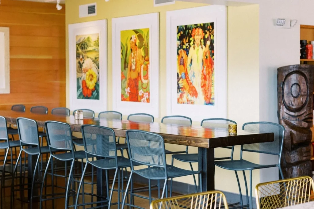 Modern restaurant interior: long wooden communal table, bar stools, and tropical art.