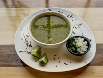 Green soup, chopped onions, herbs, lime wedges on wood.