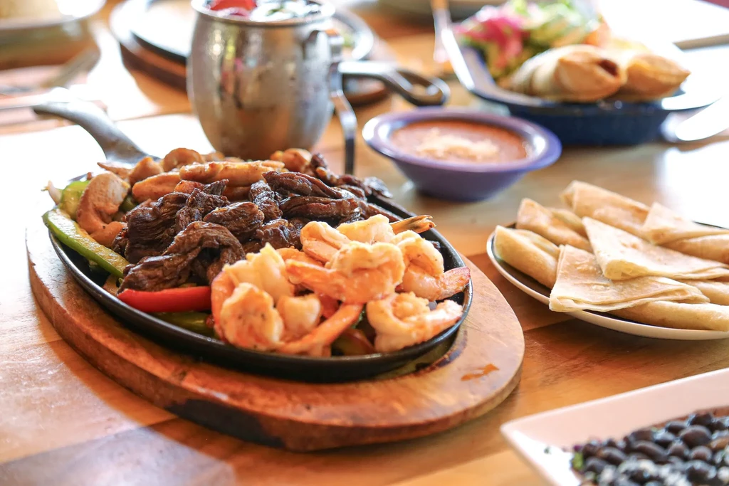 Sizzling steak, shrimp, chicken fajitas with tortillas and sides on a restaurant table.